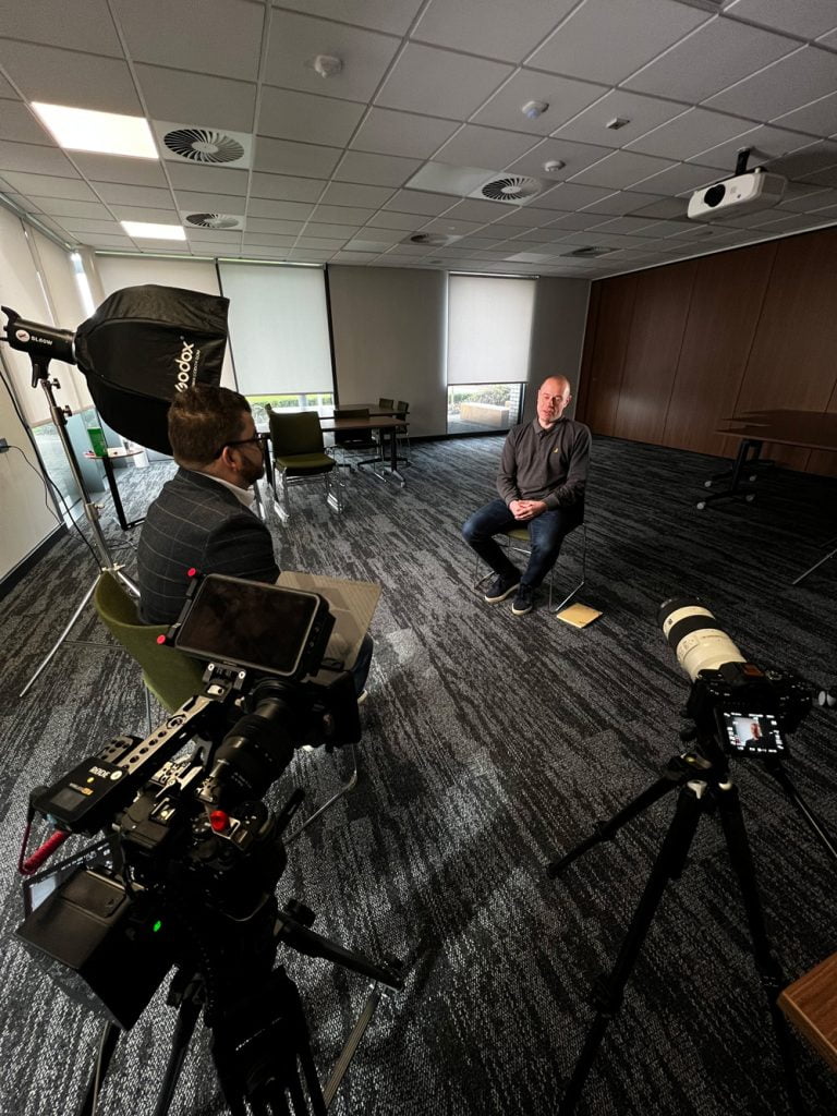 Image of two men sitting in a room filled with cameras and equipment for a video production company.